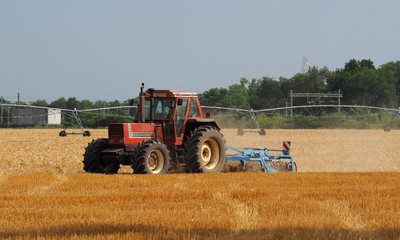 Red tractor with plough machine plows a just cut hay field. Large water irrigation system on background