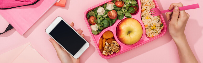 cropped view of woman holding smartphone and fork in hands near lunch box with food