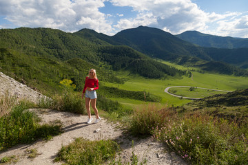 Fototapeta na wymiar A girl in a red top and blue shorts on the edge of a cliff in the Altai Mountains, below are green fields with trees and grass under a blue sky with clouds.