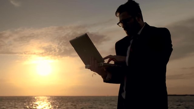 A freelance business man enthusiastically works with his laptop in the middle of the sea on the background of a magnificent sunset and kiteboarding.