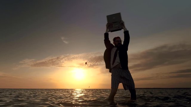 A businessman without pants being on vacation stands in the middle of the sea amid a golden sunset and rejoices at the victories achieved. A businessman enthusiastically raises up his laptop and
