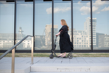 side view of an overweight woman in black dress riding on electric scooter in front of reflective windows with modern architectural buildings at Medienhafen Duesseldorf