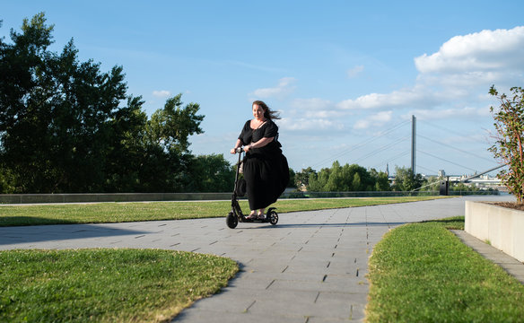 curvy overweight business woman in black dress riding an electric scooter on a sunny summer day on the sidewalk with blue cloudy sky and bridge over the rhine in the background