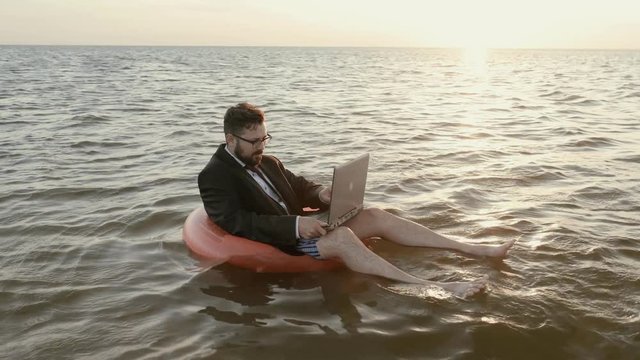 An entrepreneur sits in the middle of the sea at sunset and in a suit and no pants, in male striped underpants on a pink inflatable circle and works with a laptop.