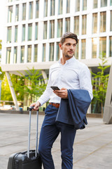 Fototapeta premium Young businessman walking outdoors on the street with suitcase using mobile phone.