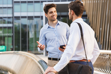 Cheerful happy young two friends colleagues business men standing near business center talking with...