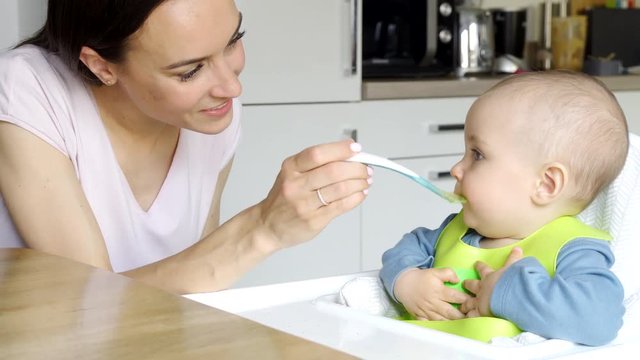 Smiling 8 Month Old Baby Boy At Home In High Chair Being Fed Solid Food By Mother With Spoon  