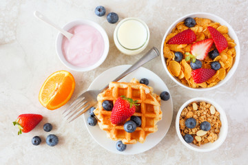 Breakfast food table scene. Fruits, cereal, waffles, yogurt and milk. Top view over a bright stone background.
