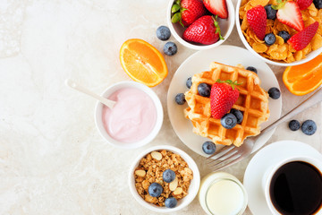 Breakfast food table scene. Fruits, cereal, waffles, yogurt and coffee. Top view over a bright stone background.