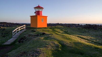 Summer Iceland - lighthouse on west cost near midnight. Low sun illuminating a red lighthouse close to rocky Atlantic shore. Green vegetation, blue sky above