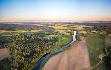 Aerial view over rural landscape in a warm summer sunset tones. Agriculture land mixing with forest and meadows. Green crop fields along the curved river. Trees creating long shadows. 