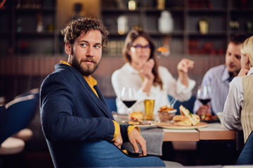 Young serious man dressed smart casual sitting at restaurant and looking over shoulder. In background his friends having dinner.