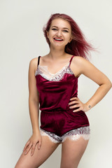 Fototapeta premium Concept portrait of a knee-high pretty sexy girl with purple hair in beautiful red lingerie on a white background. She smiles, happy with life, happy. Hair flying, front view. Made in a studio.