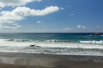 Panoramic view of Aimasiga beach with volcanic black sand and lone rocks sticking out of the sea foam on the north coast of the island Tenerife, Spain..Sunny day, blue sky.