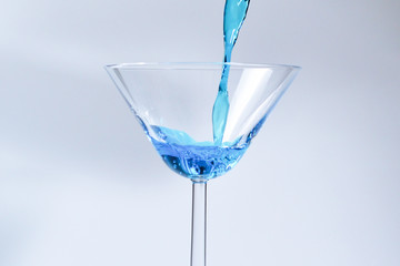 Cocktail with blue liquid in glass. Glass with blue water pouring with liquid with splashes and drops. Martini glass filling with alcohol with splashes on white background. Refreshing drink concept.
