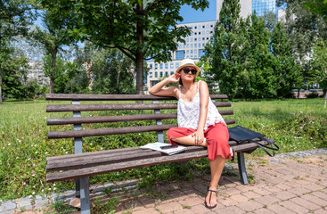 Beautiful young positive and independent French hipster tourist girl with hat and sunglasses adventurous world traveler sitting on the bench in city on a new trip to a new country and new adventure