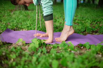 Thin brunette girl plays sports and performs yoga poses in a summer park. Green forest on the background. Woman doing exercises on the yoga mat, hands and feet close-up