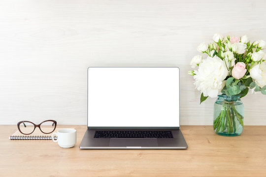Office desk table with laptop computer with blank screen mockup and flowers bouquet, coffee cup, glasses, notepad on wooden surface. Minimal style feminine workplace concept.