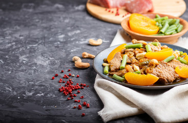 Fried pork with peaches, cashew and green beans on a black concrete background, side view. raw meat, red pepper, linen textile.