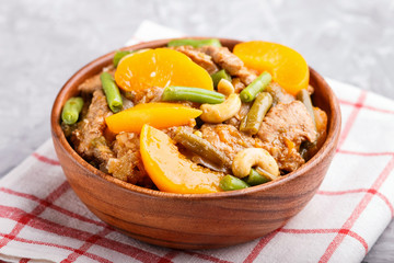 Fried pork with peaches, cashew and green beans in a wooden bowl on a gray concrete background, close up.