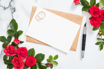 Mockup wedding invitation card or love letter with craft paper envelope, rose flowers, bridal rings on marble background. Minimal flat lay style composition, top view, overhead.