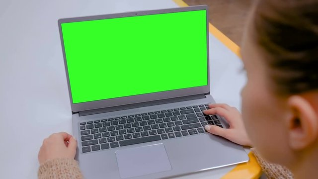 Woman sitting at table and looking at grey laptop computer device with blank green screen in cafe, home or office. Technology, chroma key, template, mockup and entertainment concept