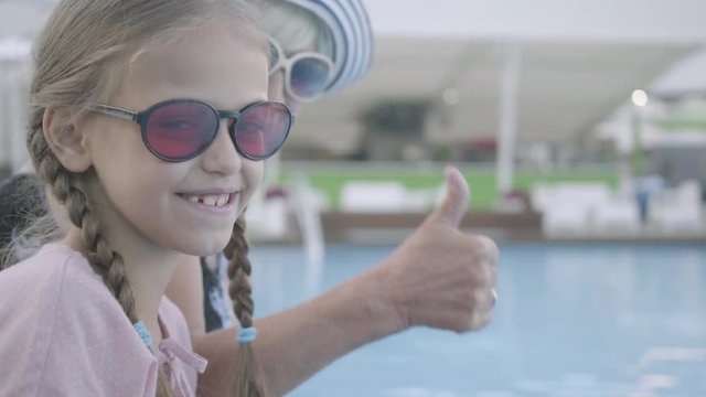 Little funny girl with pigtails and mature woman sitting on the edge of the pool smiling in the camera and showing thumb up. Happy friendly family. Rest in hotel