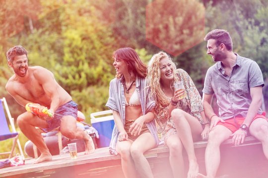 Cheerful Shirtless Man Spraying Water With Squirt Gun On Friends Sitting At Lakeshore