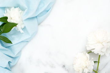 Top view frame of white peony flowers and pastel blue cloth on marble background. Minimal flat lay style composition, beauty or fashion blog hero banner template.