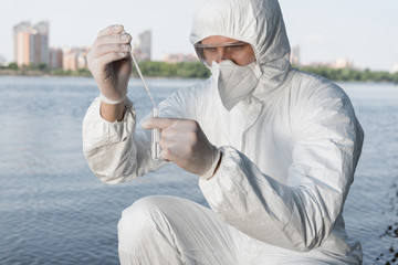 water inspector in protective costume and respirator taking water sample at river