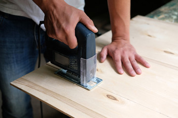 Carpenter is sawing a plywood sheet with electric jig saw machine in carpentry workshop. Close up hand with jigsaw. Carpentry concept.