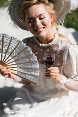 happy young victorian woman with closed eyes holding fan and wine glass