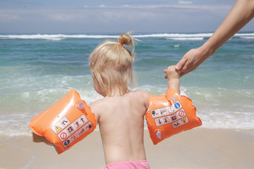 Candid portrait of a happy toddler enjoying a day at the beach. Girl running to the sea wearing inflatable armbands, back view