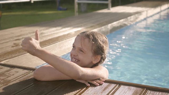 Cute funny girl with pigtails laughing, showing thumb up looking out of the pool, holding on to the edge. Preteen child having fun in the hotel complex