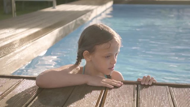 Adorable funny girl with pigtails laughing, showing thumb up looking out of the pool, holding on to the edge. Preteen child having fun in the hotel complex