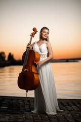 girl in white beautiful dress with a cello stands by the lake at sunset