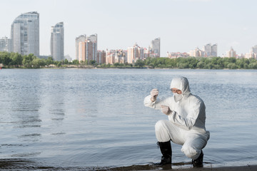 water inspector in protective costume and respirator taking water sample at river