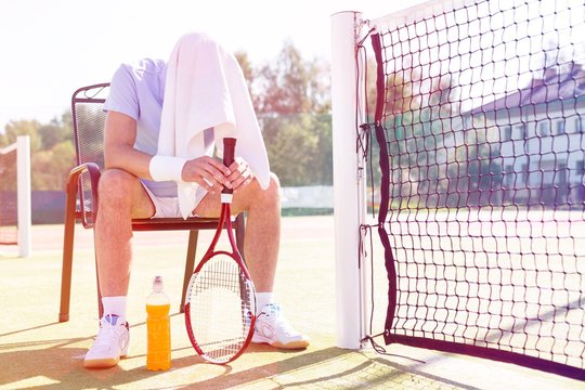 Full length of tired mature man with covered head sitting on chair by net at tennis court on sunny day - Powered by Adobe