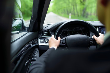 View from a back seat in a car, on a driver hands on a wheel. View of road and trees during trip.