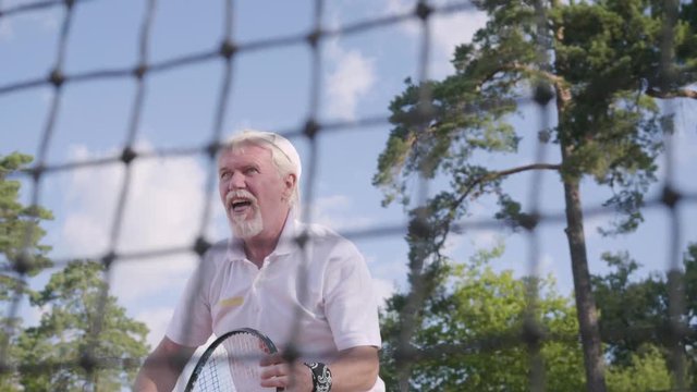 Portrait Joyful happy smiling mature man playing tennis on the tennis court. The old man throws the ball with the racket. Recreation and leisure outdoors.