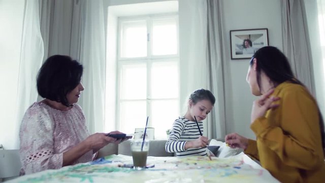 A small girl with mother and grandmother sitting at table at home, painting pictures.
