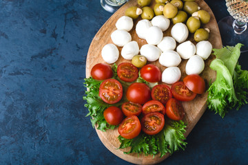 summer snack of mozzarella, tomatoes, olives and greens on wooden board. Dark blue background. Horizontal, place for text