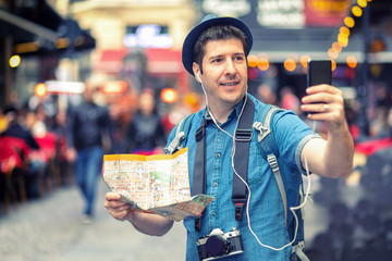 Smiling man tourist holding city map having roaming video call on mobile phone on European crowded streets full of pubs  - obrazy, fototapety, plakaty