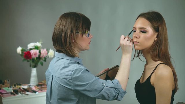 Makeup artist lifts girl's chin and paints her eyes using a palette with shadows. Person is smart and has a model appearance. On the background is wedding bouquet of peonies and roses in awesome vase.
