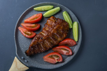 baked pork ribs with honey lie with fresh sliced ​​tomatoes and cucumbers on a dark gray concrete tabletop