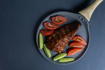 baked pork ribs with honey lie with fresh sliced ​​tomatoes and cucumbers on a dark gray concrete tabletop