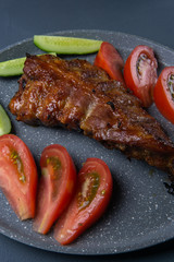 baked pork ribs with honey lie with fresh sliced ​​tomatoes and cucumbers on a dark gray concrete tabletop