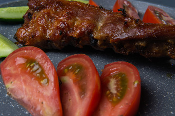 baked pork ribs with honey lie with fresh sliced ​​tomatoes and cucumbers on a dark gray concrete tabletop