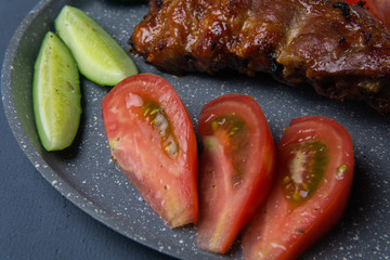 baked pork ribs with honey lie with fresh sliced ​​tomatoes and cucumbers on a dark gray concrete tabletop