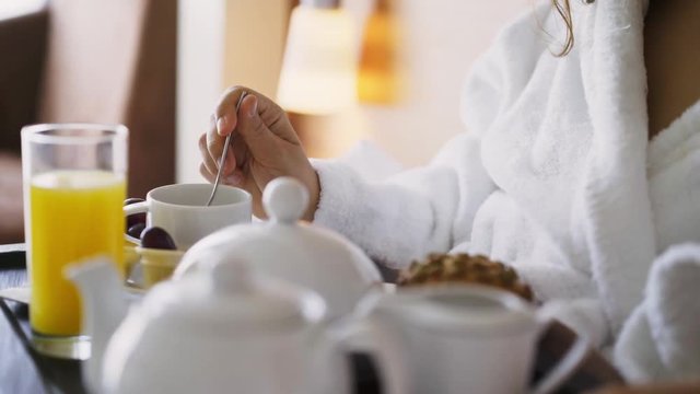 Girl enjoying morning in light hotel apartment with full length windows. Woman in white bathrobe having breakfast in bed, detail view tray full of food and female hand stirring spoon in cup 